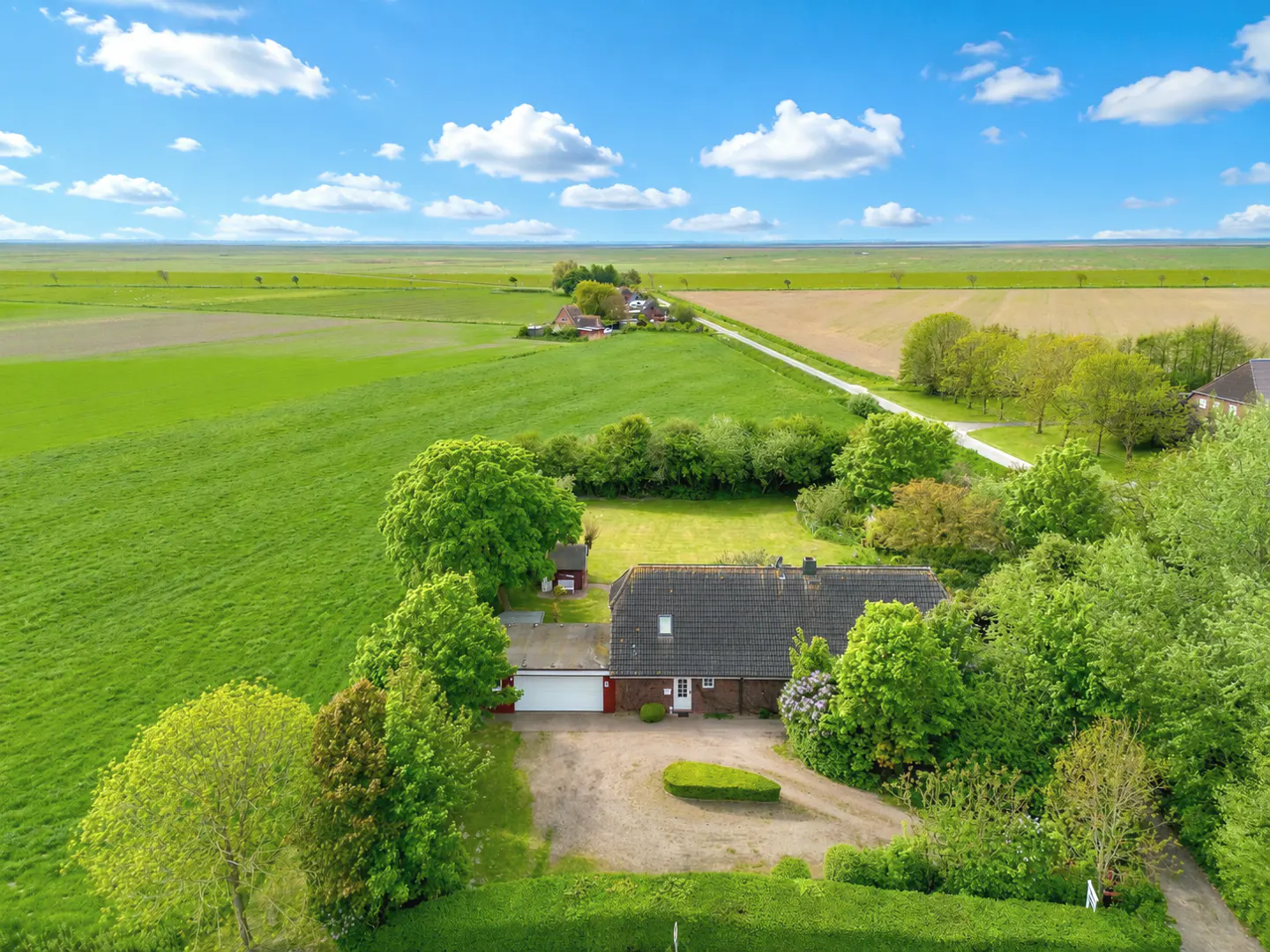 Aerial view of a house with a gray roof, surrounded by green trees and fields under a blue sky with white clouds.