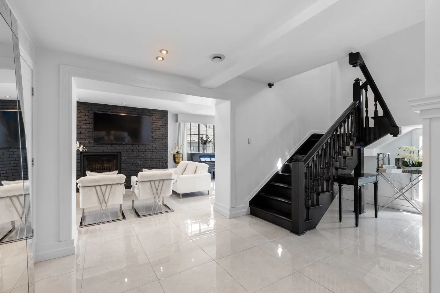 Bright foyer with white tile floors, black staircase, and living room view. Living room has a black brick fireplace and white furniture.