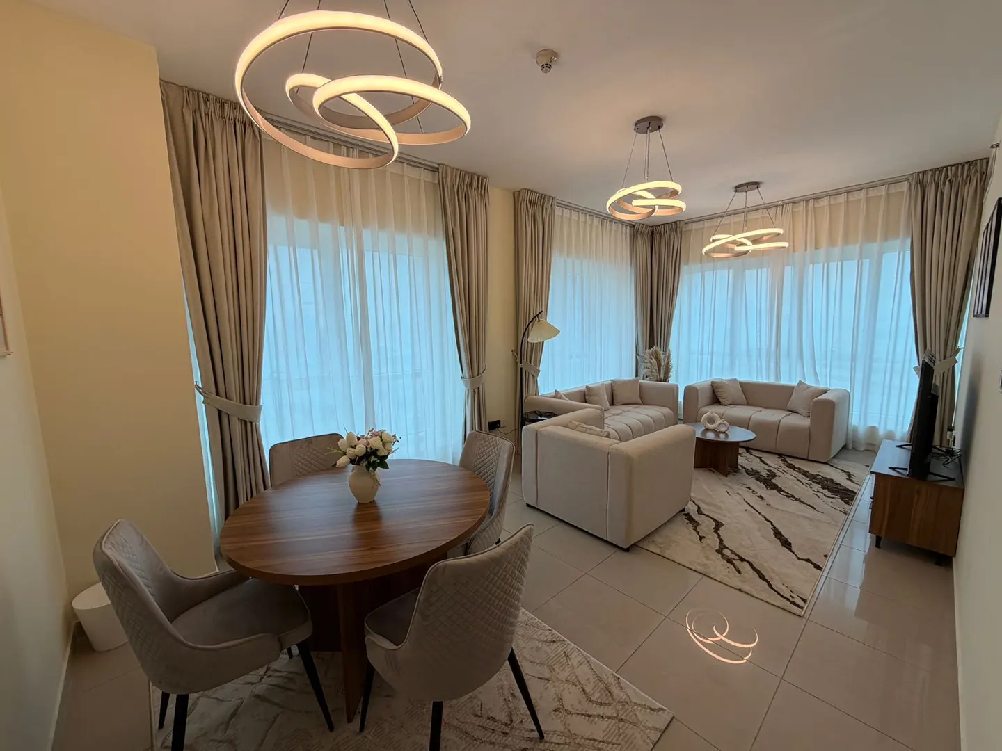 Bright, modern apartment with a round wood dining table, four chairs, and a living room with beige sofas and a patterned rug.