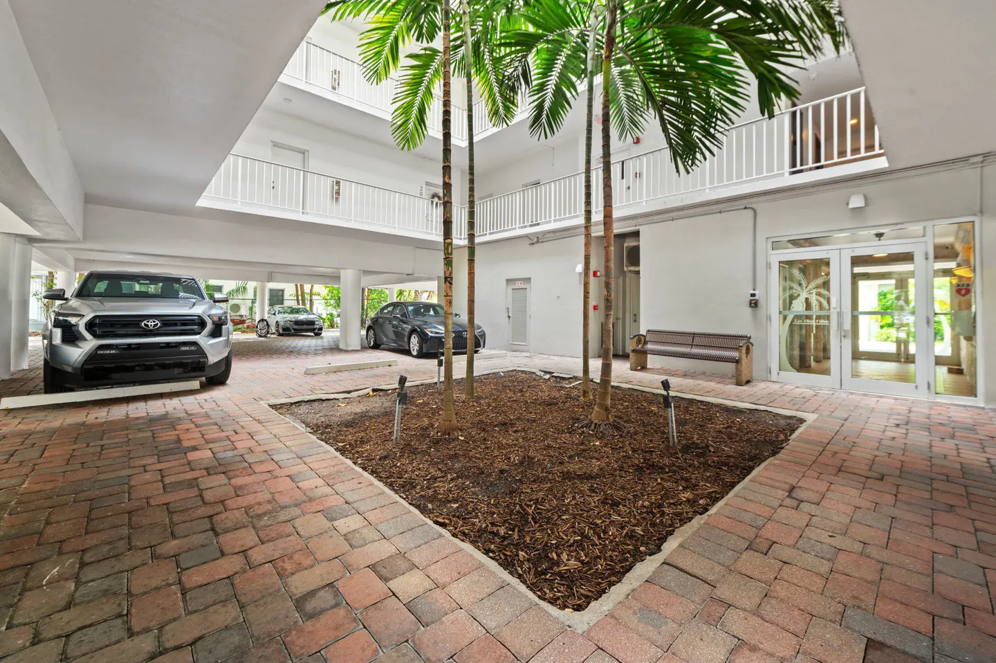 Courtyard view of a white building with palm trees, brick pavers, and parked cars. A bench sits near the entrance.