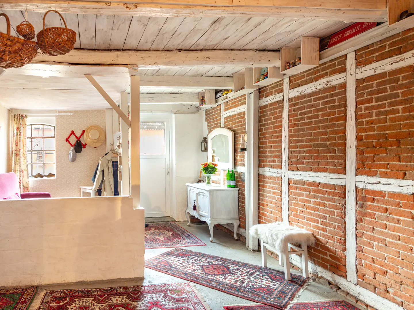 Interior view of a rustic home with exposed brick and white wood beams. Baskets hang from the ceiling, and oriental rugs cover the floor. A white dresser and bench add charm.