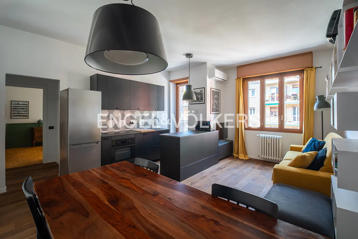 Interior view of a modern apartment with a wooden dining table, black kitchen cabinets, and a yellow sofa near a window.