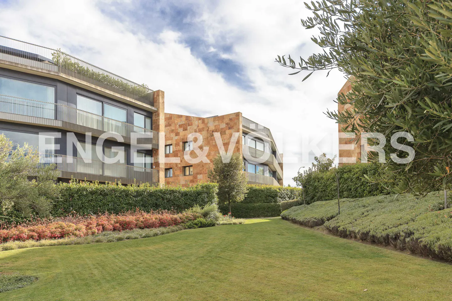 Modern apartment building with stone and glass facade, surrounded by green lawn and manicured hedges. Engel & Volkers logo visible.