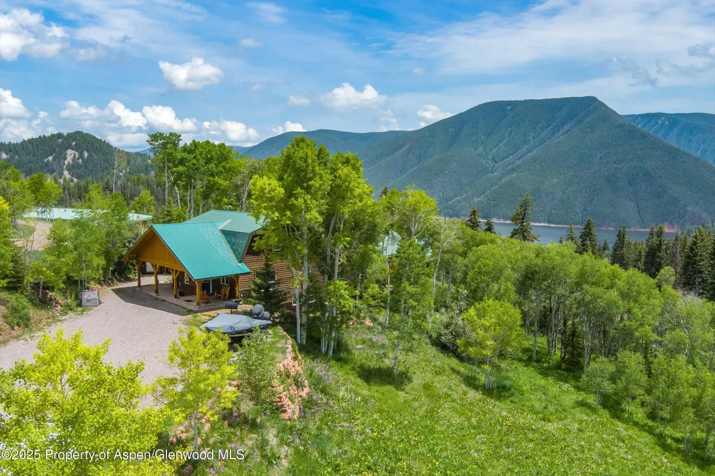 Aerial view of a log cabin with a green roof, surrounded by trees and mountains under a blue sky.