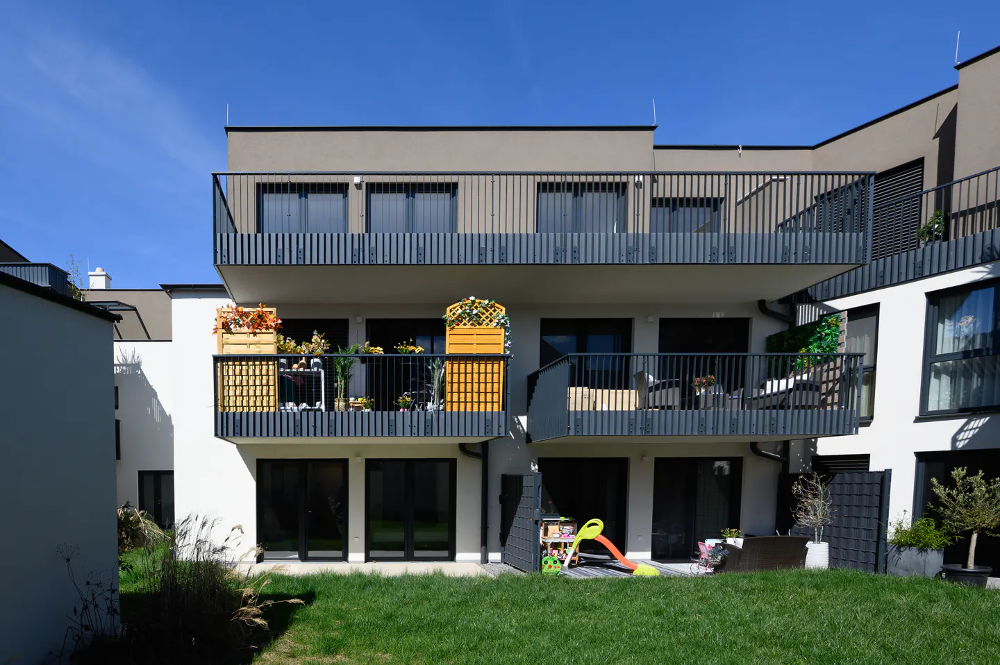 Modern apartment building with balconies, white walls, and a green lawn. Balconies have flowers and plants. A children's slide is visible on the ground floor.