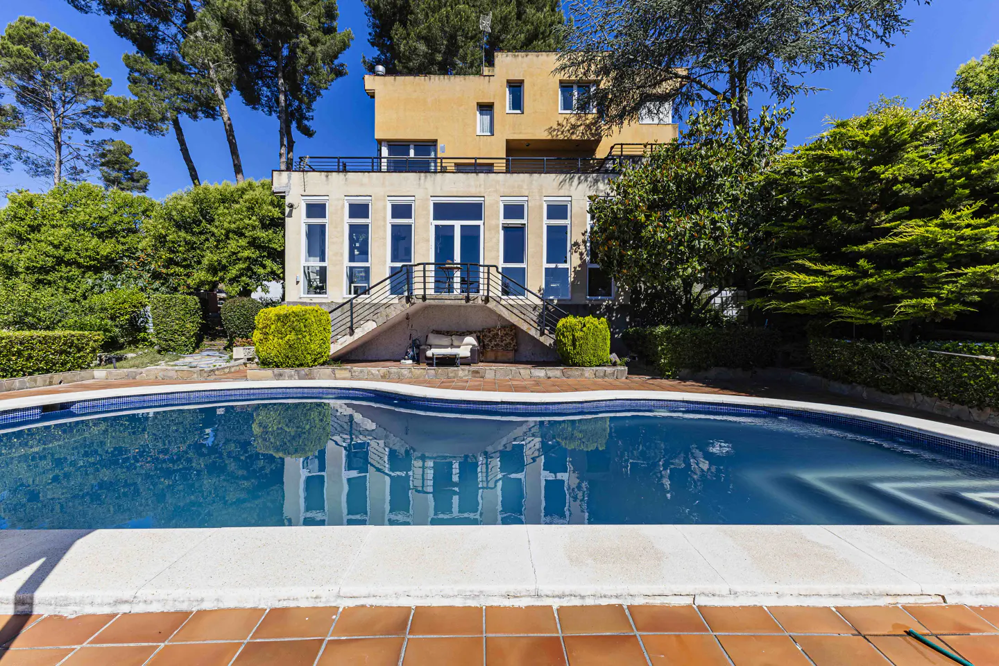 A two-story tan house with a swimming pool in the foreground, surrounded by lush green trees under a clear blue sky.