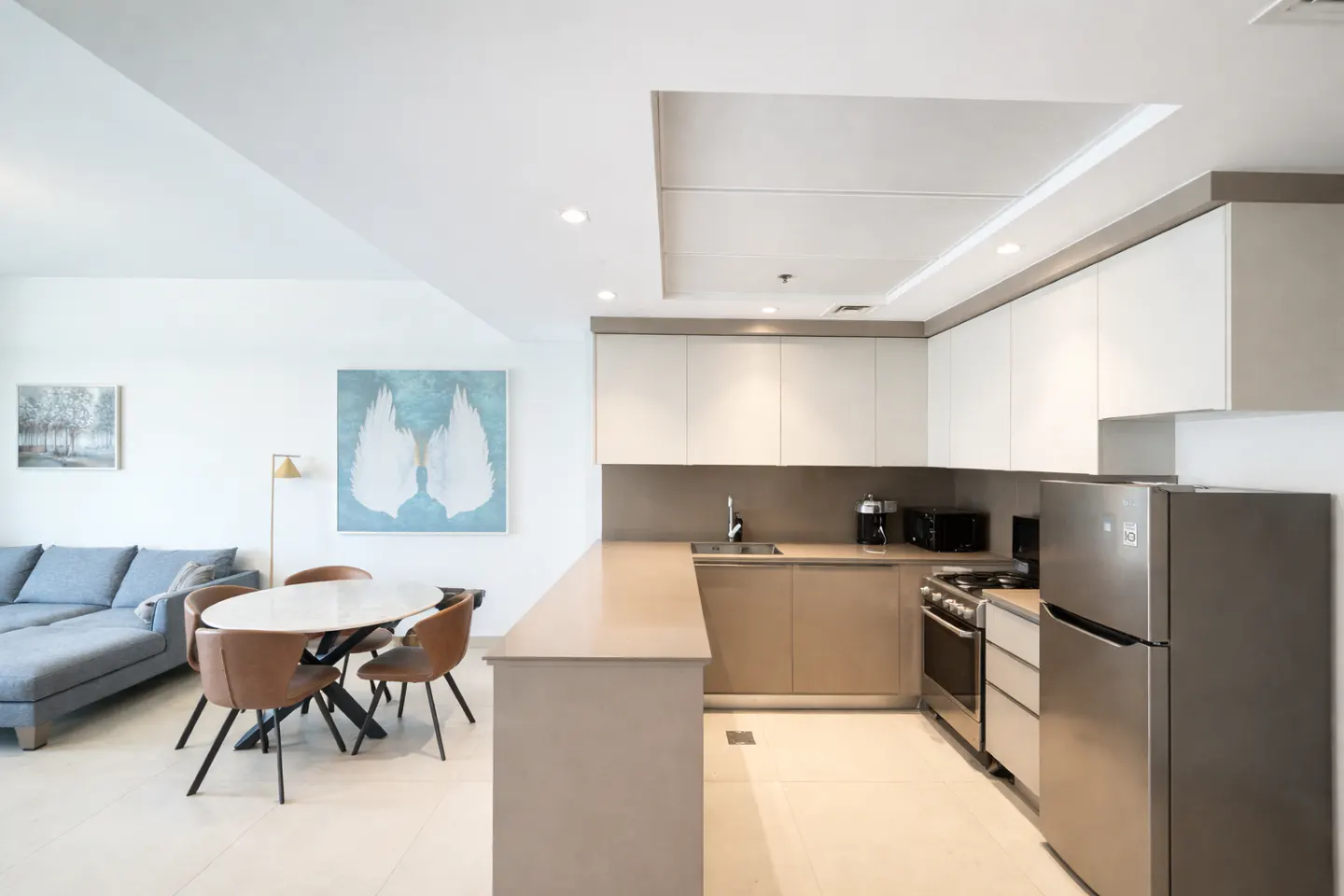 A bright, modern kitchen and dining area with white cabinets, stainless steel appliances, and a round table with brown chairs. A blue sofa is visible in the background.