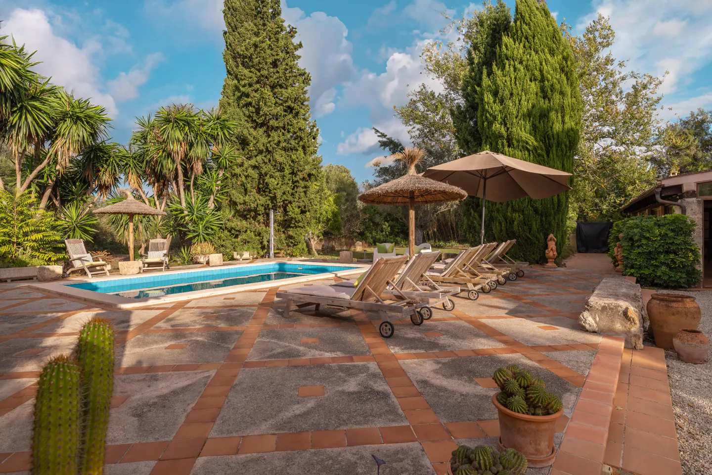 Outdoor pool area with lounge chairs, umbrellas, and lush greenery. The pool is rectangular and blue. The patio is tiled with red accents.