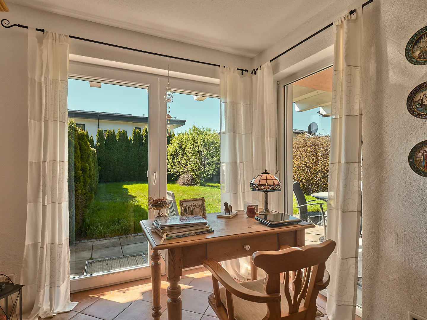 A bright sunroom features a wooden desk and chair, with a view of a green lawn and garden through glass doors.