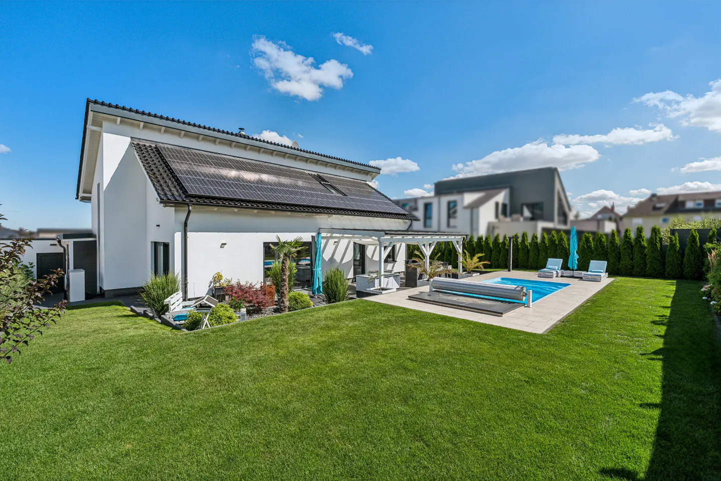 Backyard view of a modern white house with solar panels, a pool, a pergola, and a green lawn under a blue sky.