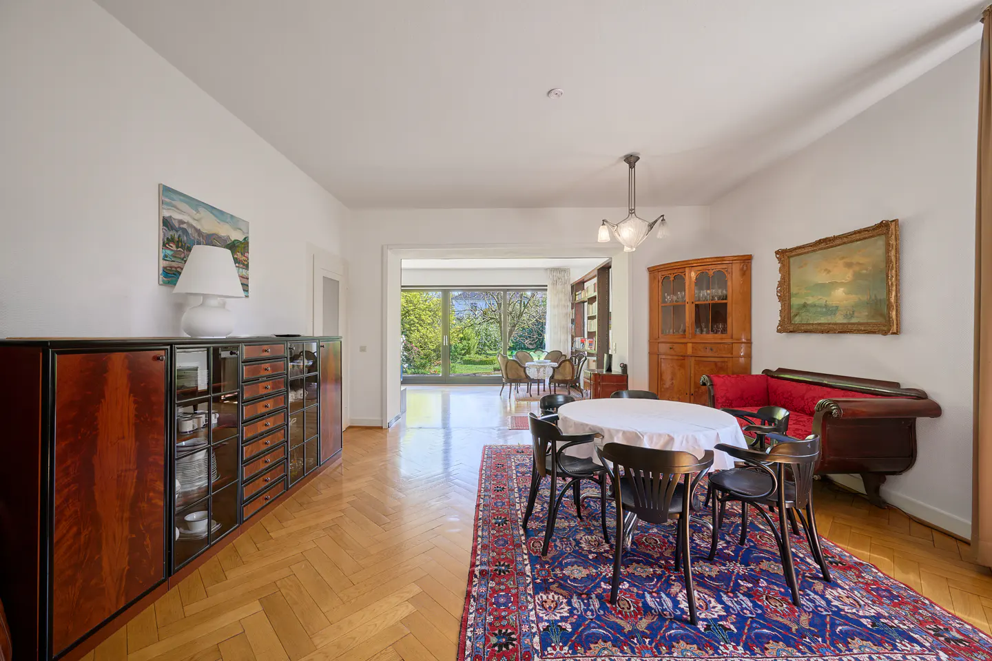 Bright dining room with wood floors, white walls, and a blue rug. A round table with black chairs sits on the rug. A large window looks out to a green yard.