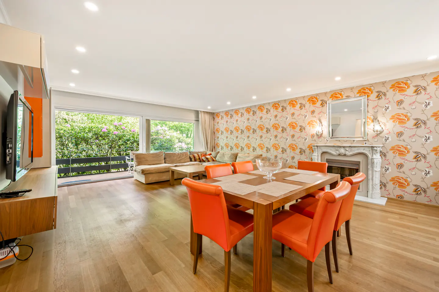 Bright living room with hardwood floors, orange chairs around a wood table, and floral wallpaper. A fireplace and sofa are in the background.