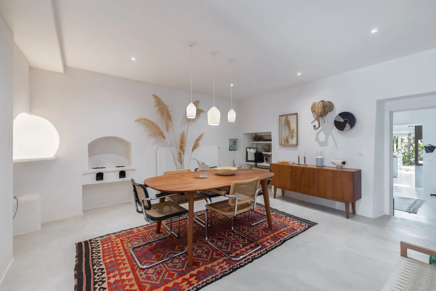 Bright dining room with white walls, a wooden table, and four chairs on a red patterned rug. A wooden sideboard and decorative items adorn the walls.