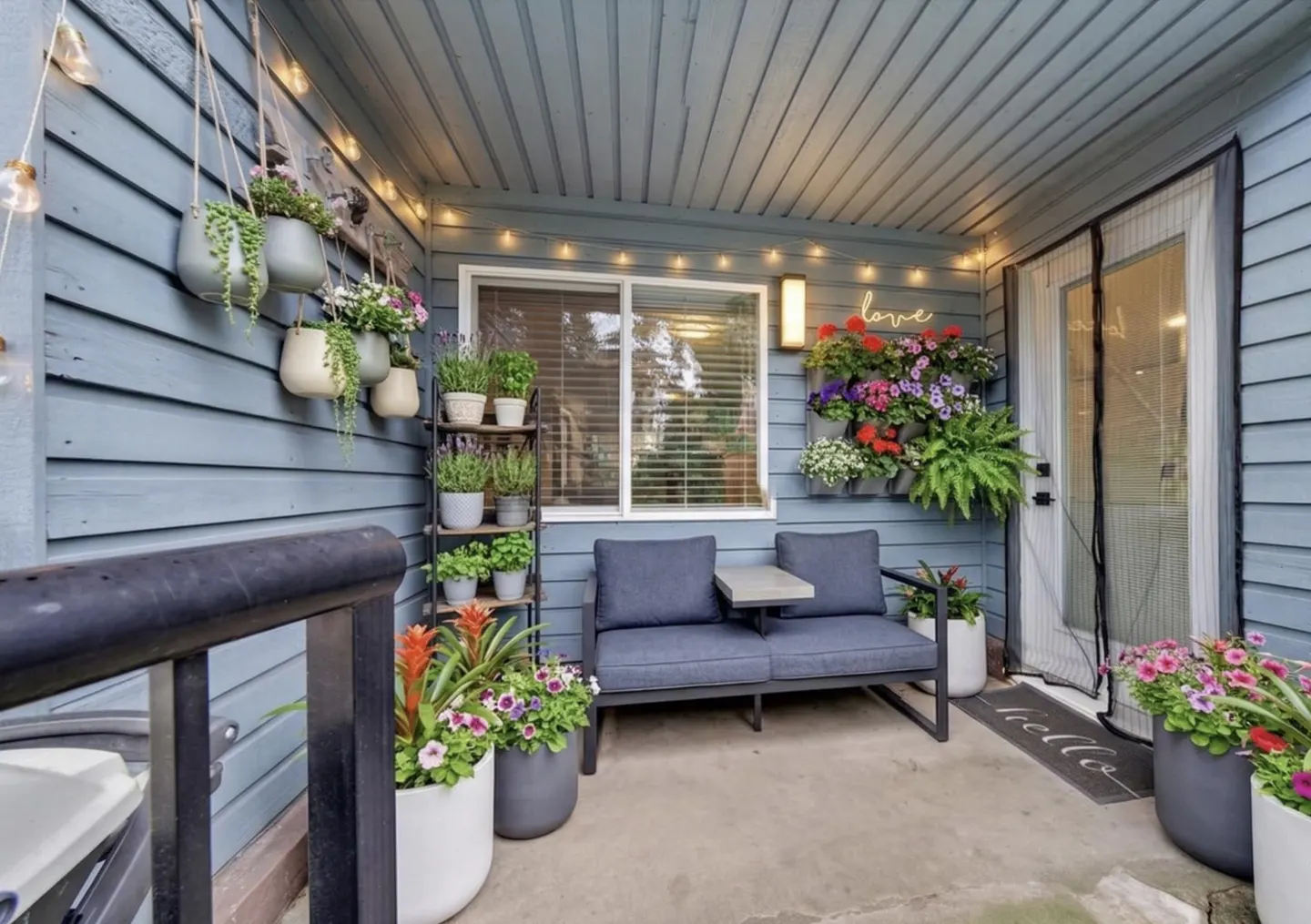Cozy blue porch with string lights, potted plants, and a loveseat. A "love" sign and "hello" mat add charm.