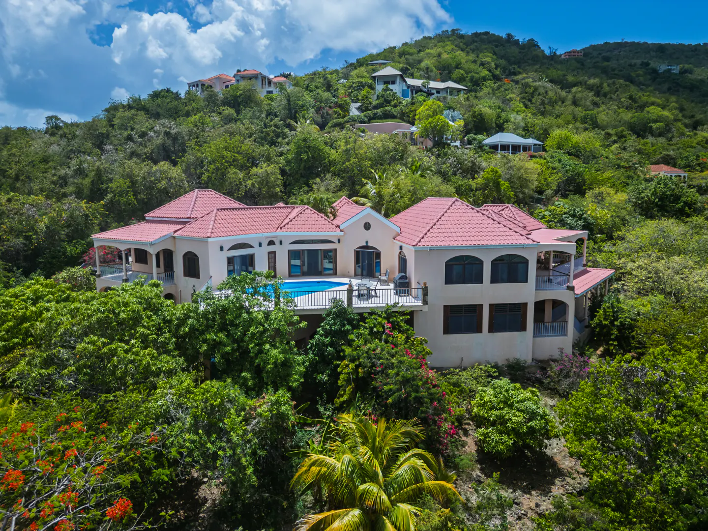 Aerial view of a beige villa with a red tile roof and swimming pool, nestled in a lush, green tropical hillside.