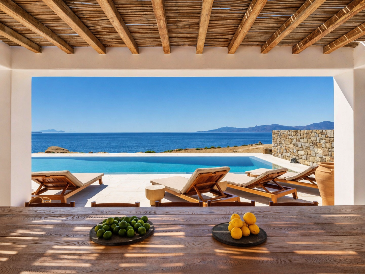 View from a shaded patio with a wooden table, limes, lemons, pool, lounge chairs, and ocean view.