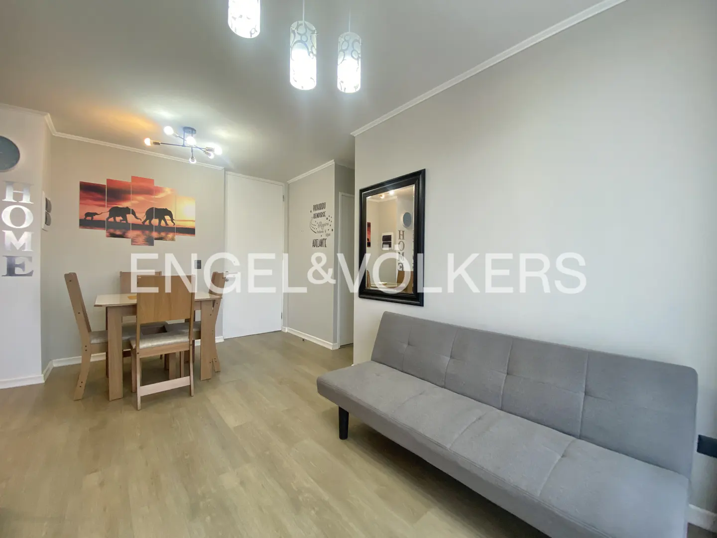 A bright living room with a gray sofa, a wooden dining table, and an elephant-themed wall art. The Engel & Volkers logo is superimposed.