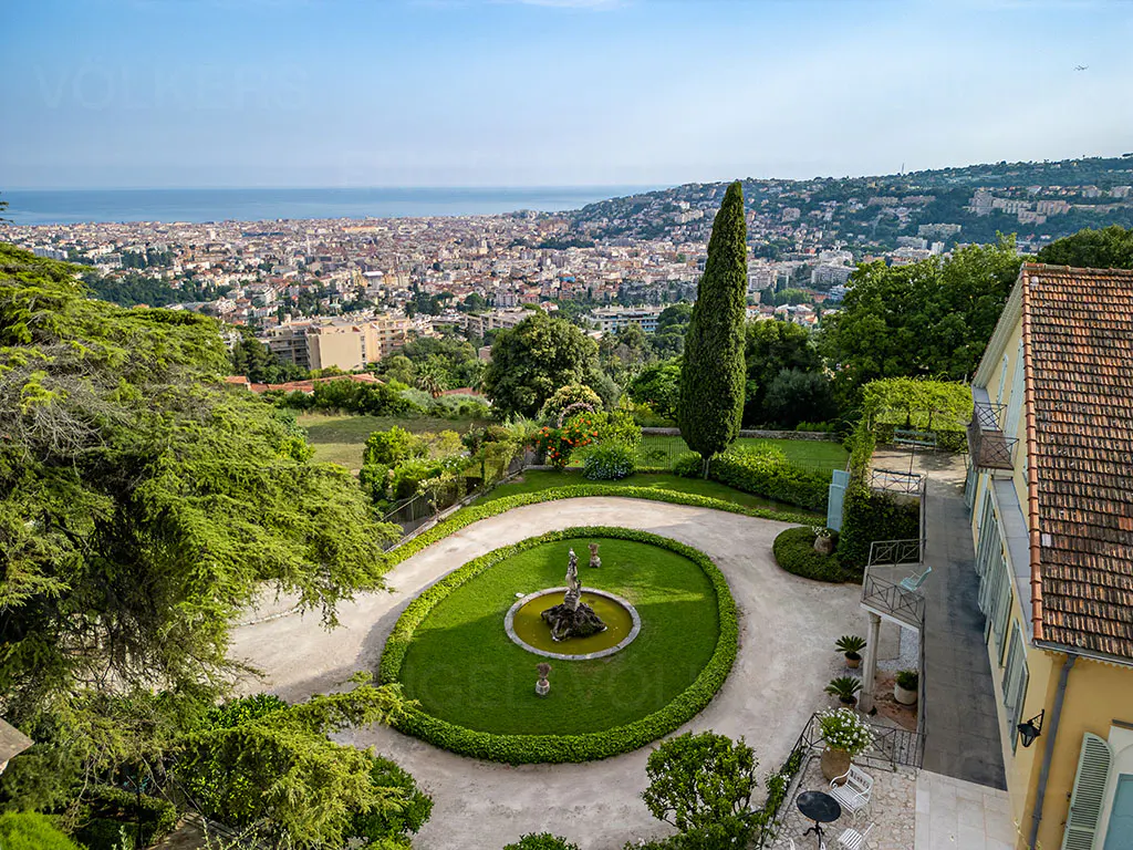 Aerial view of a property with a fountain, garden, and city view. The house has a red tile roof and yellow walls.