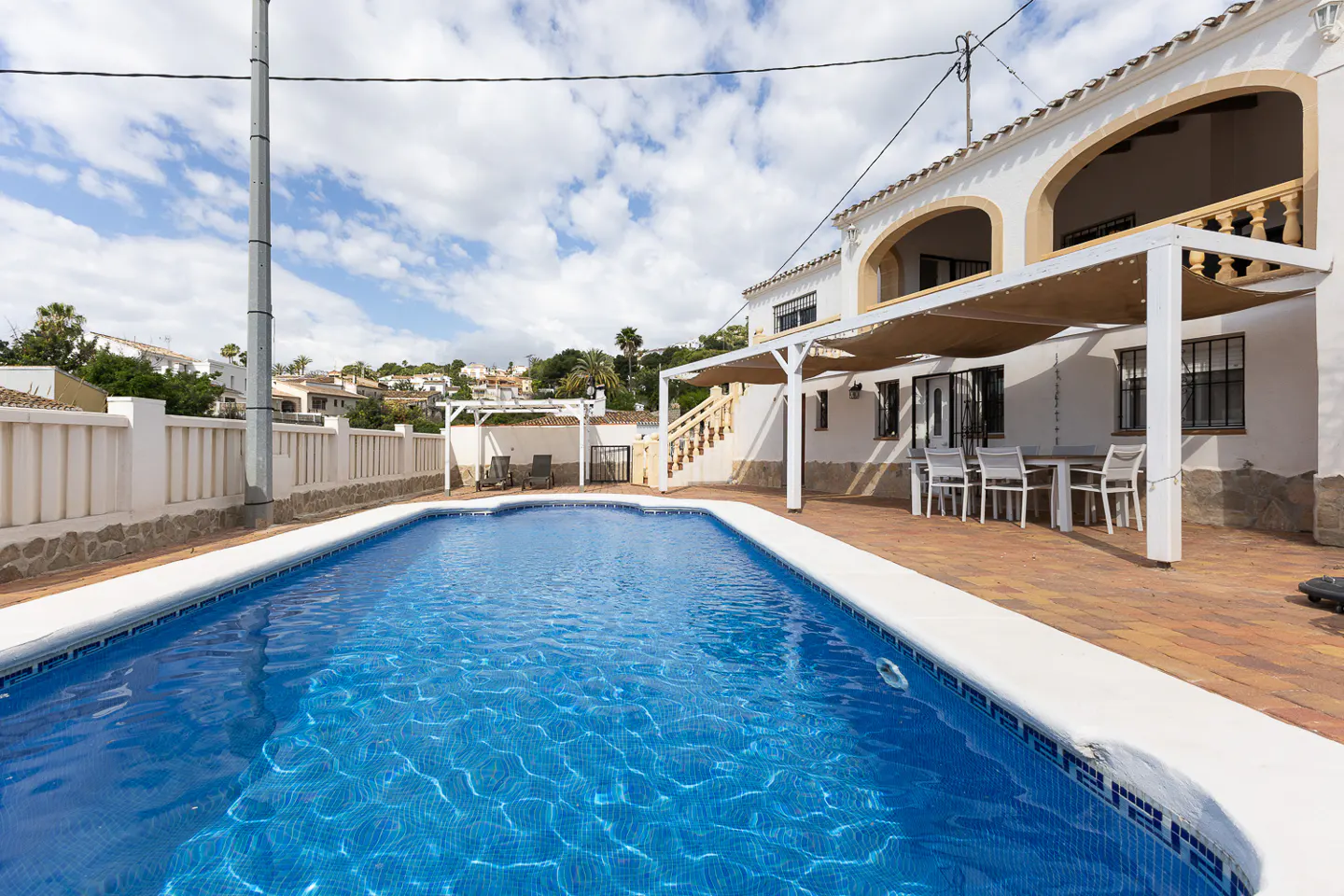 A sunny backyard with a blue tiled pool, white fence, and a two-story white house with arched balconies. A patio with a dining set is visible.