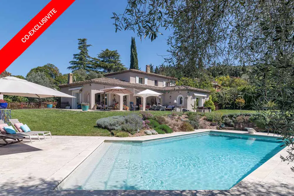 Exterior view of a two-story beige house with a pool, lawn, and patio furniture on a sunny day. "Co-Exclusive" banner in red.