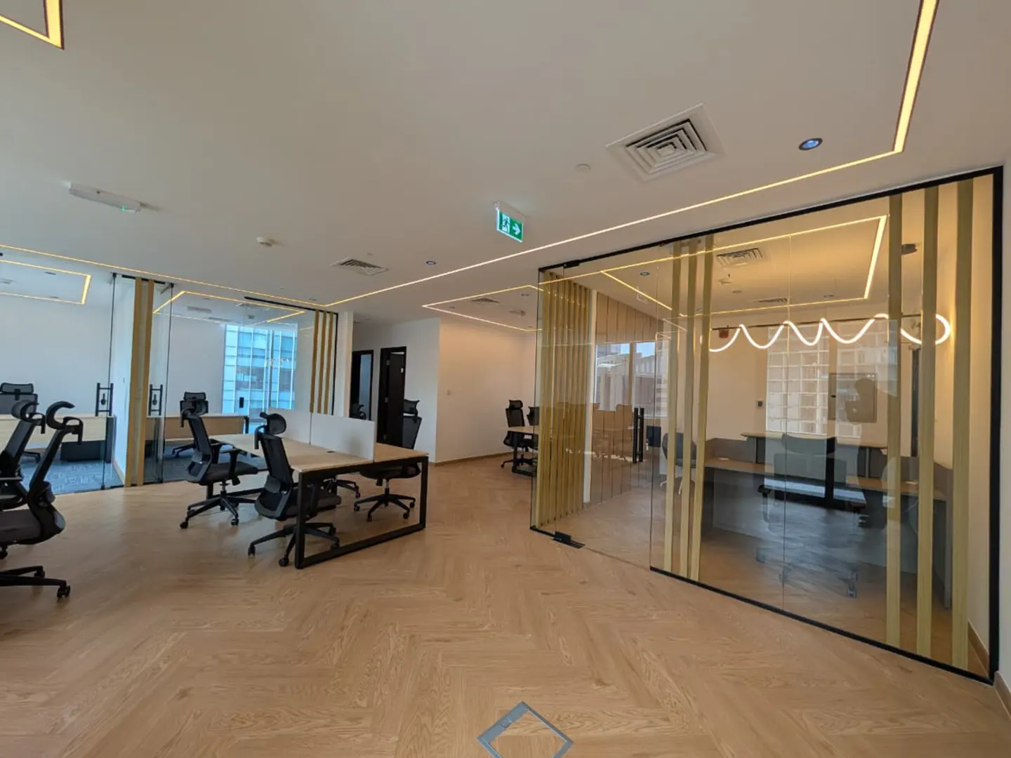 A modern office space with desks, black chairs, and glass-enclosed rooms. The floor is light wood, and the ceiling has recessed lighting.
