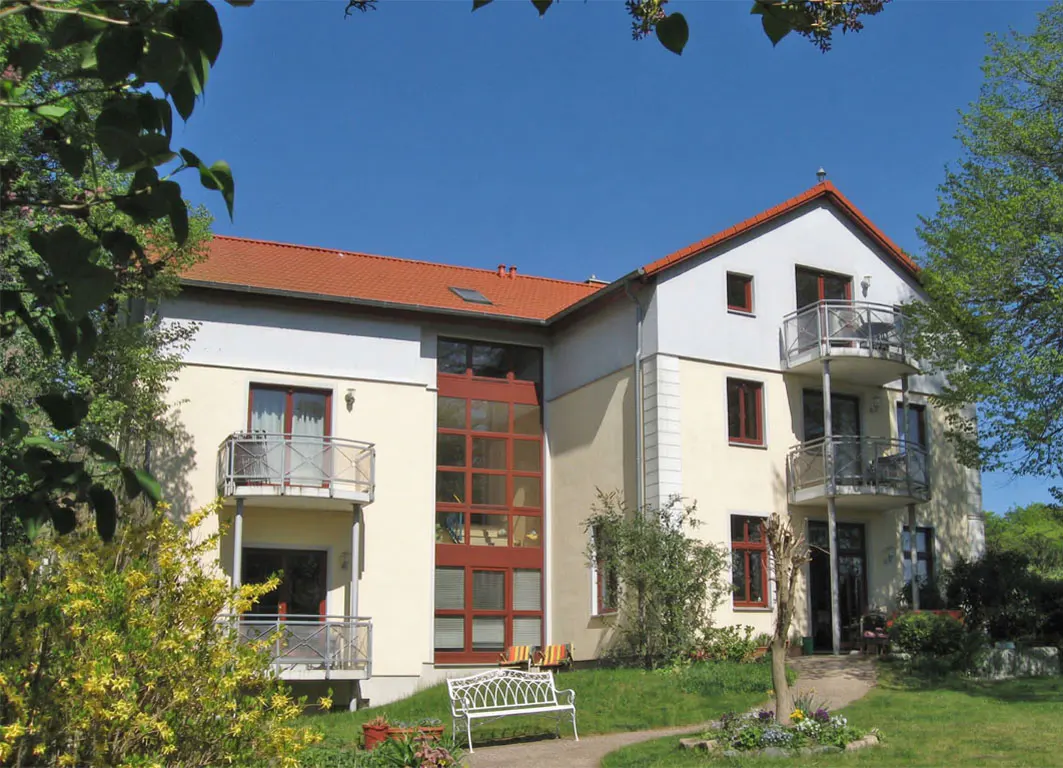 Exterior view of a two-story building with balconies, a red tile roof, and a white bench in the front yard.