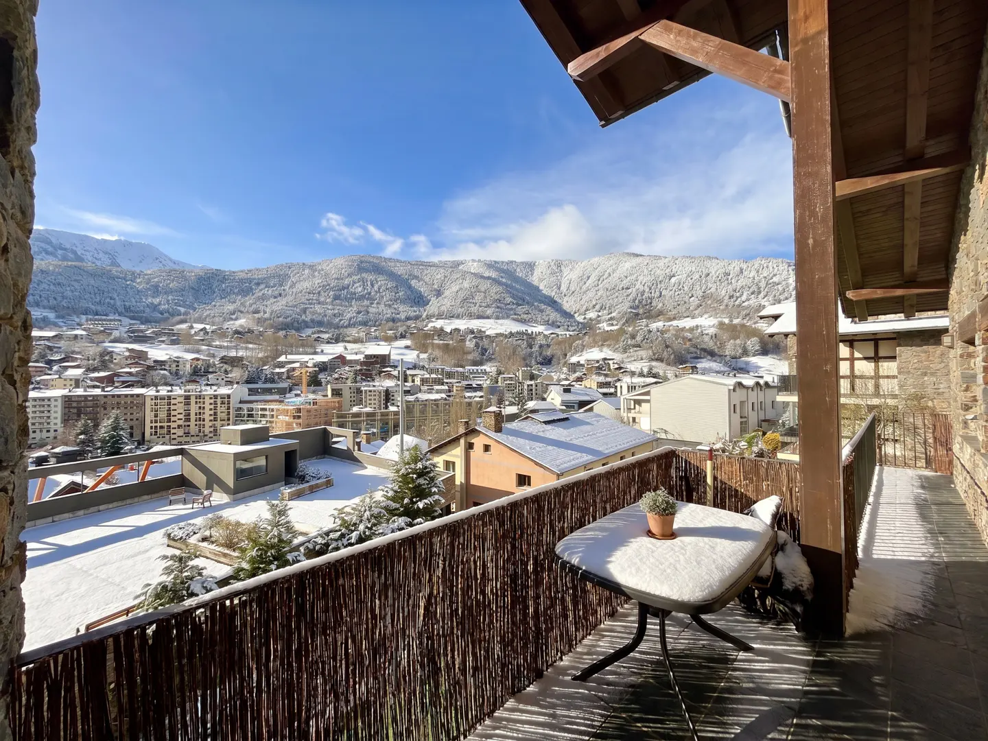 Balcony view of a snow-covered town and mountains under a blue sky. A table with a potted plant sits on the balcony.