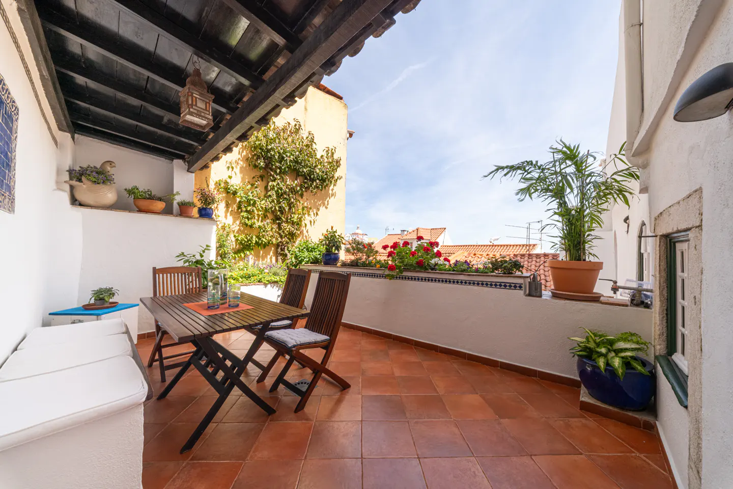 Outdoor patio with terracotta tile, table, chairs, and white cushioned bench. Plants and flowers line the balcony overlooking red rooftops.
