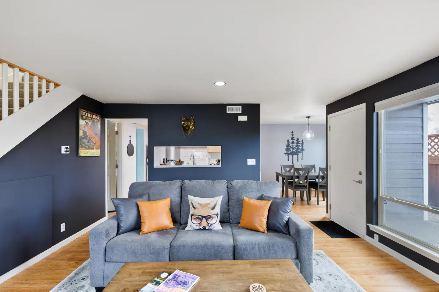 Living room with gray sofa, orange pillows, and fox decor. Dark blue walls, wood floors, and a dining area visible in the background.