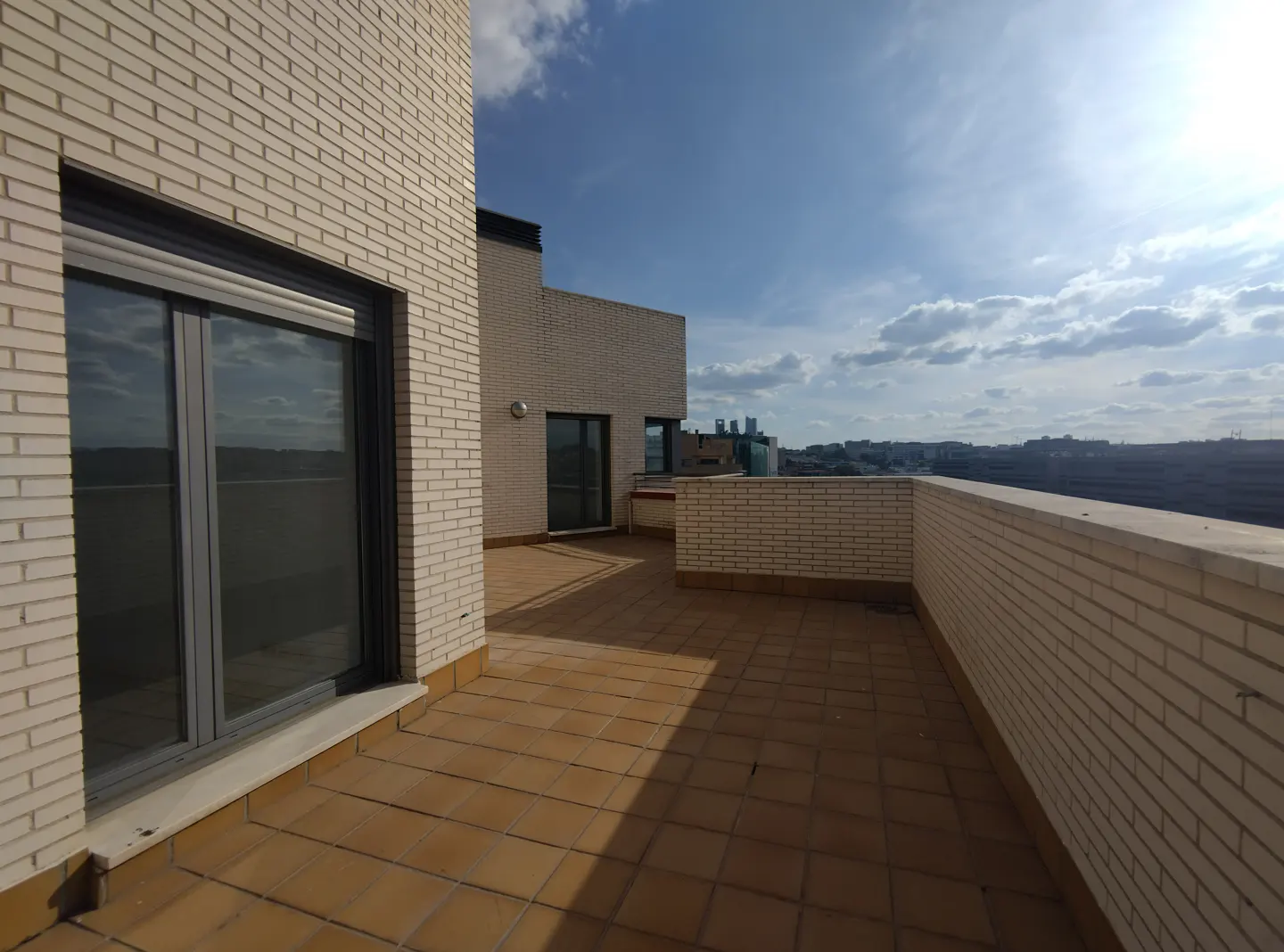 A rooftop terrace with tan brick walls and tile flooring under a blue sky with clouds.