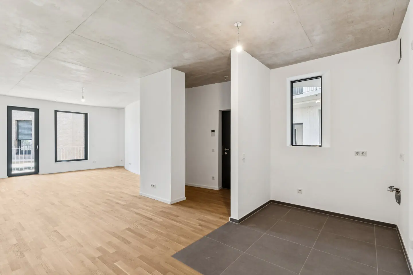 Unfurnished apartment with hardwood floors, white walls, and a concrete ceiling. Black framed windows and a black door are visible.