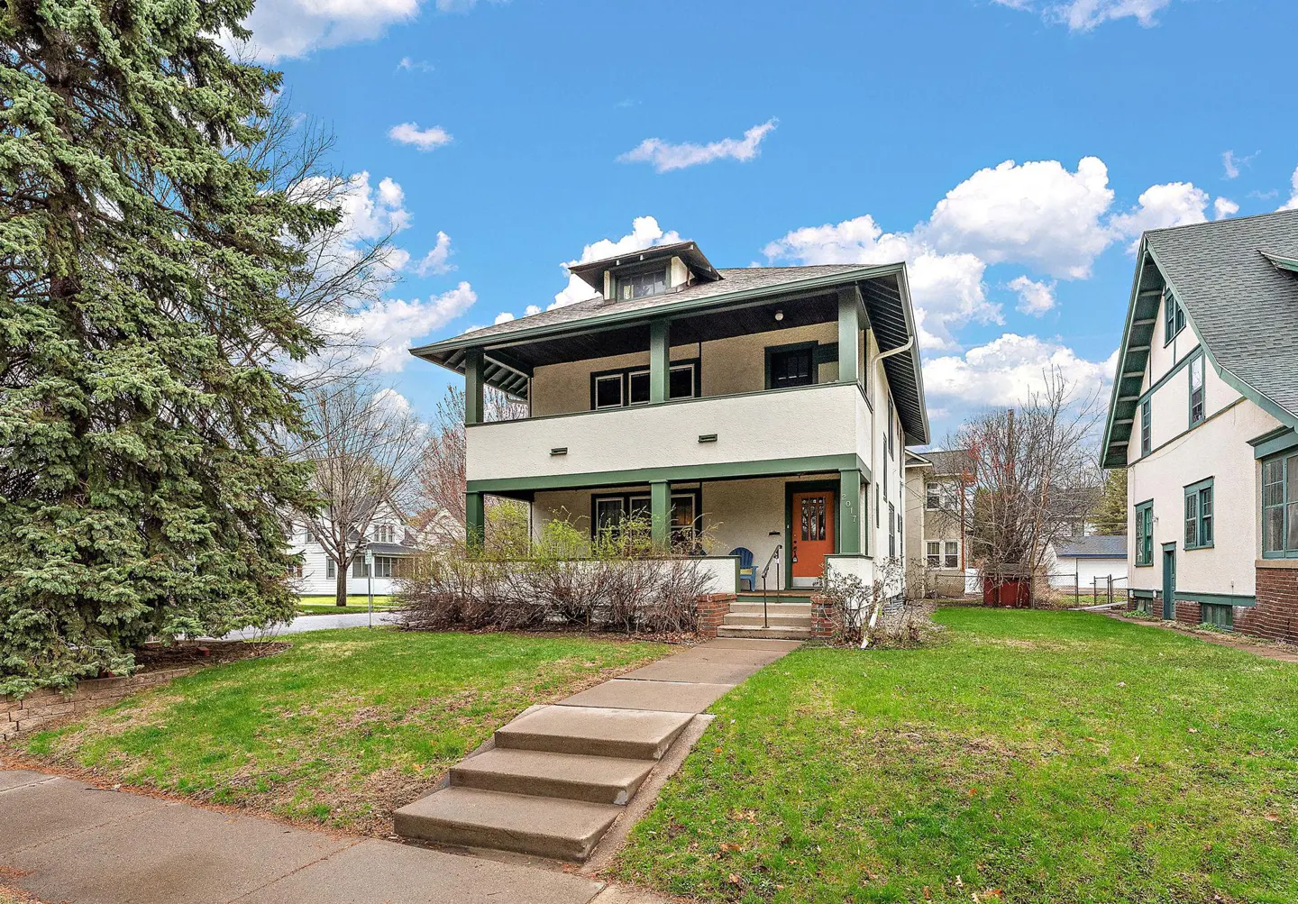 Two-story house with a porch, green trim, and an orange door, set on a green lawn under a blue sky.