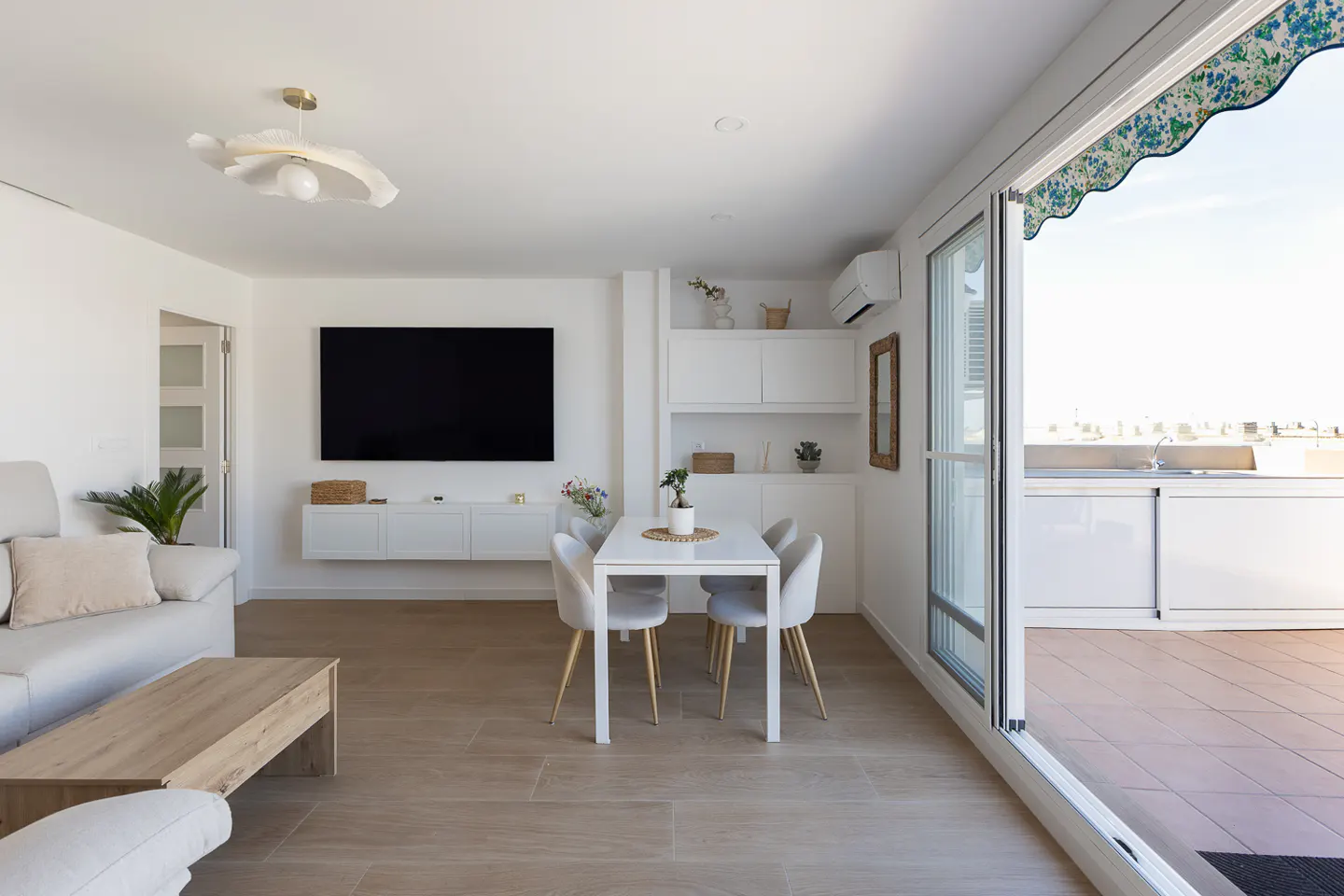 Bright, modern living room with white walls, wood floors, and a large TV. A dining table sits near sliding glass doors to a balcony.