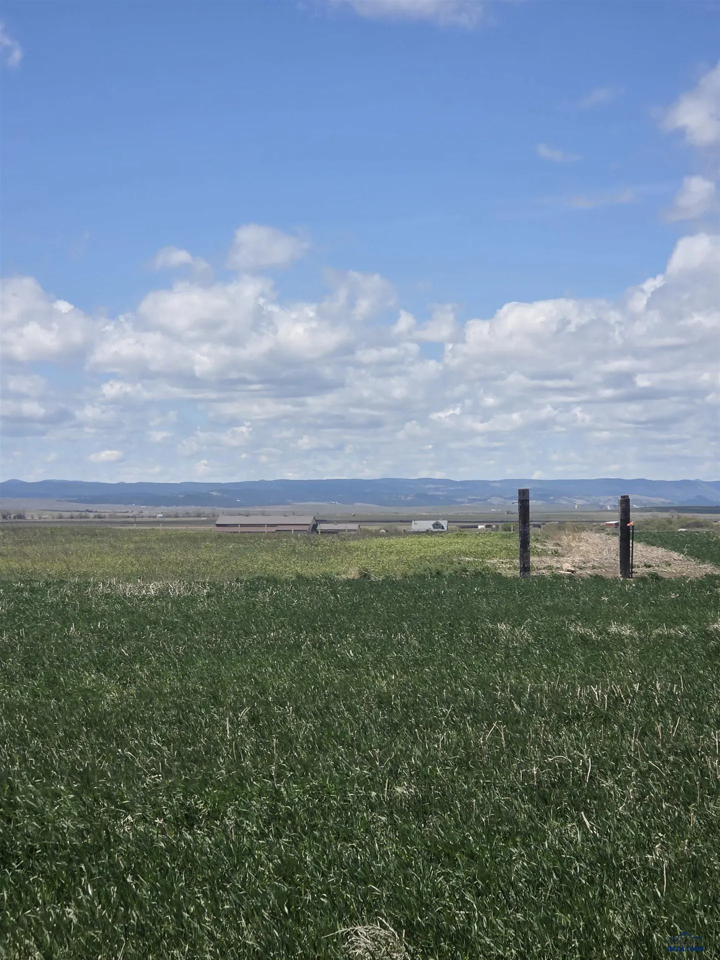 Green field under a blue, cloudy sky. Two dark posts mark a break in the field. Buildings are visible in the distance.