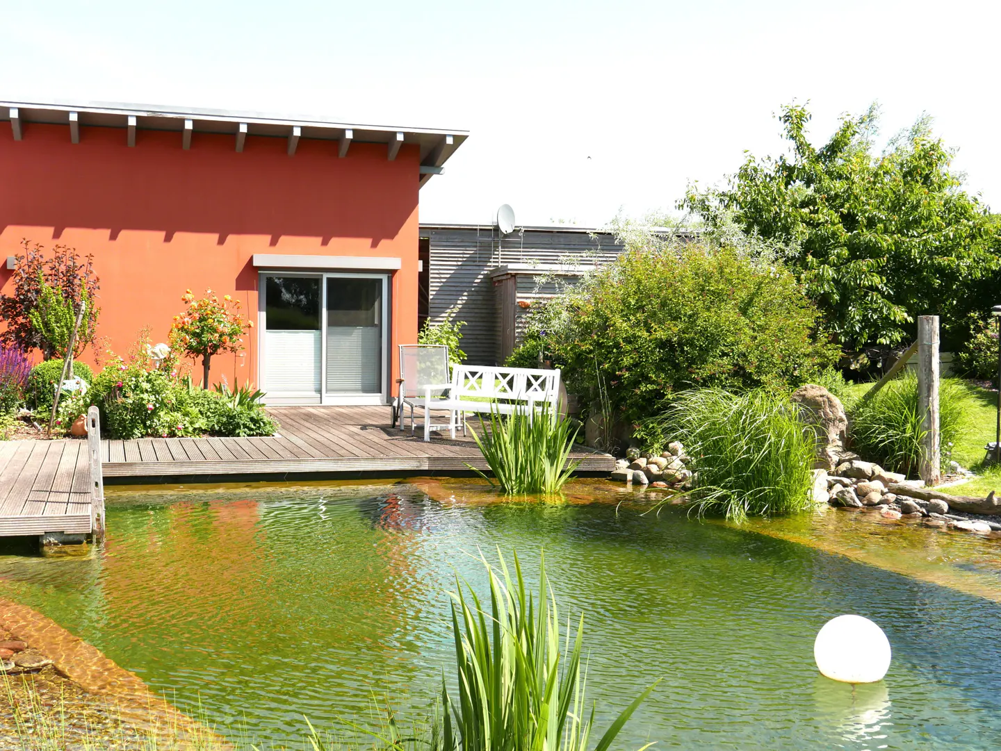 Exterior view of a red house with a pond, wooden deck, white bench, and lush greenery.