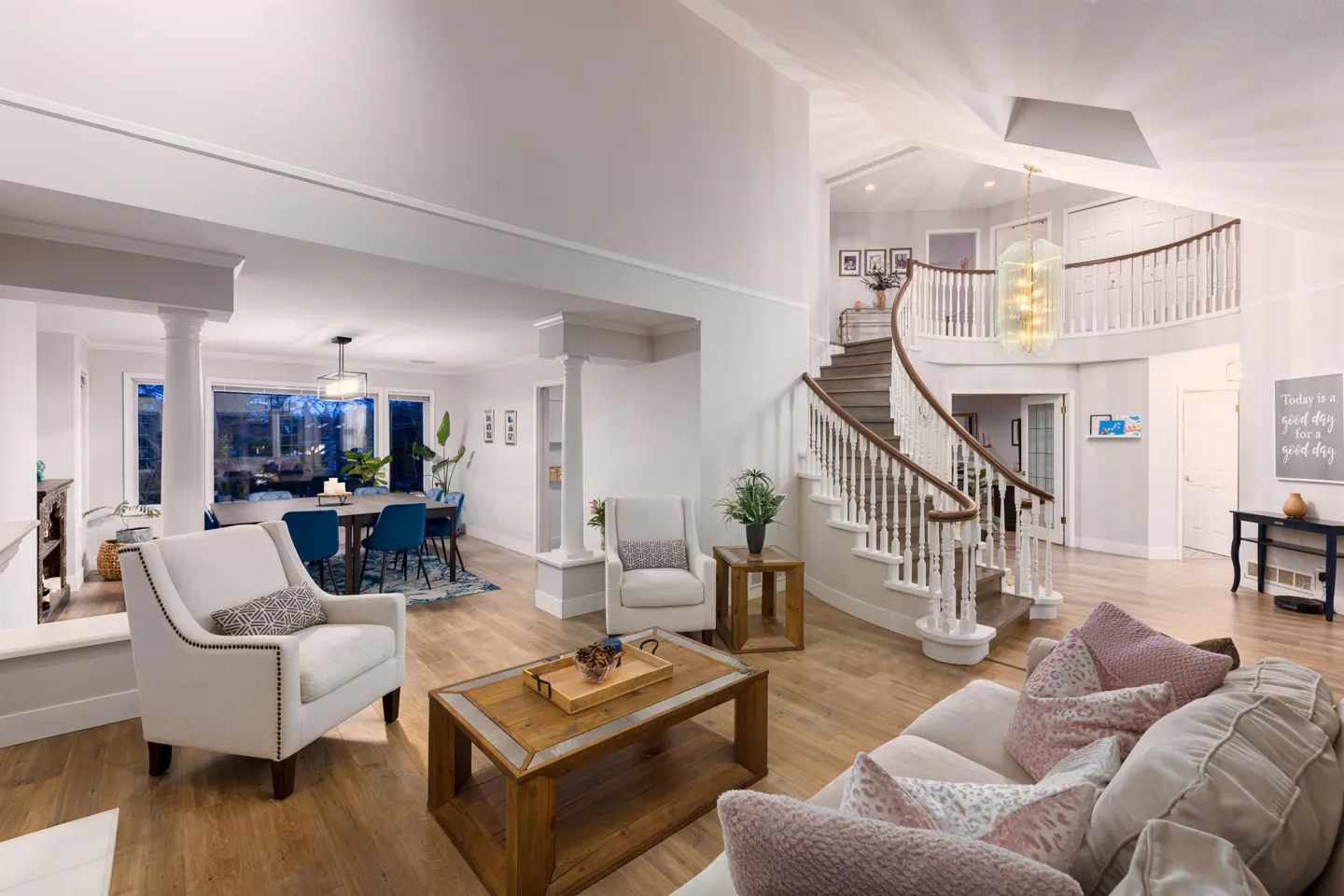 Open-concept living room with hardwood floors, white walls, and a winding staircase. Dining area with blue chairs visible. Natural light.