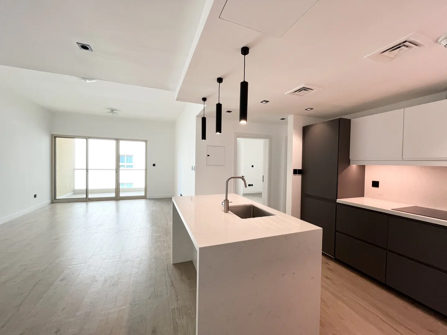 A bright, modern apartment interior with a white kitchen island, dark cabinets, and a living area with sliding glass doors.