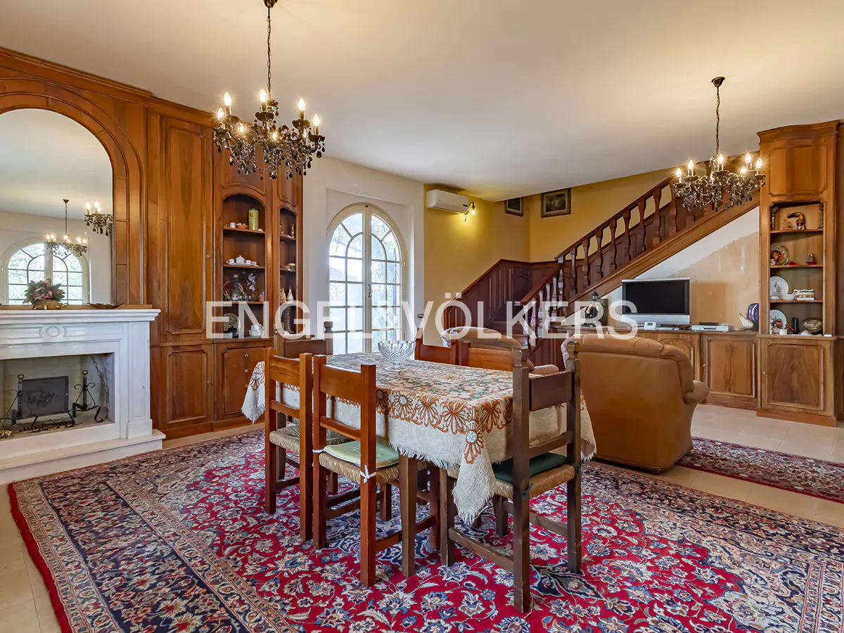 A dining room with wooden furniture, a fireplace, and a red patterned rug. Chandeliers hang from the ceiling. A staircase is visible in the background.