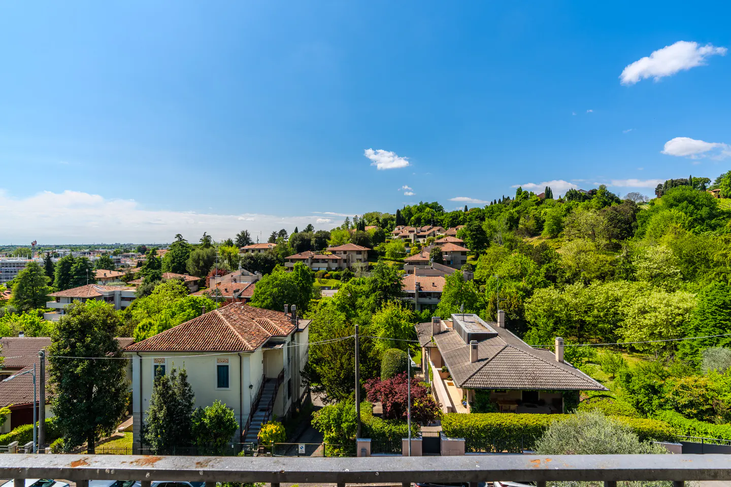 View of a hillside neighborhood with houses and lush green trees under a blue sky.
