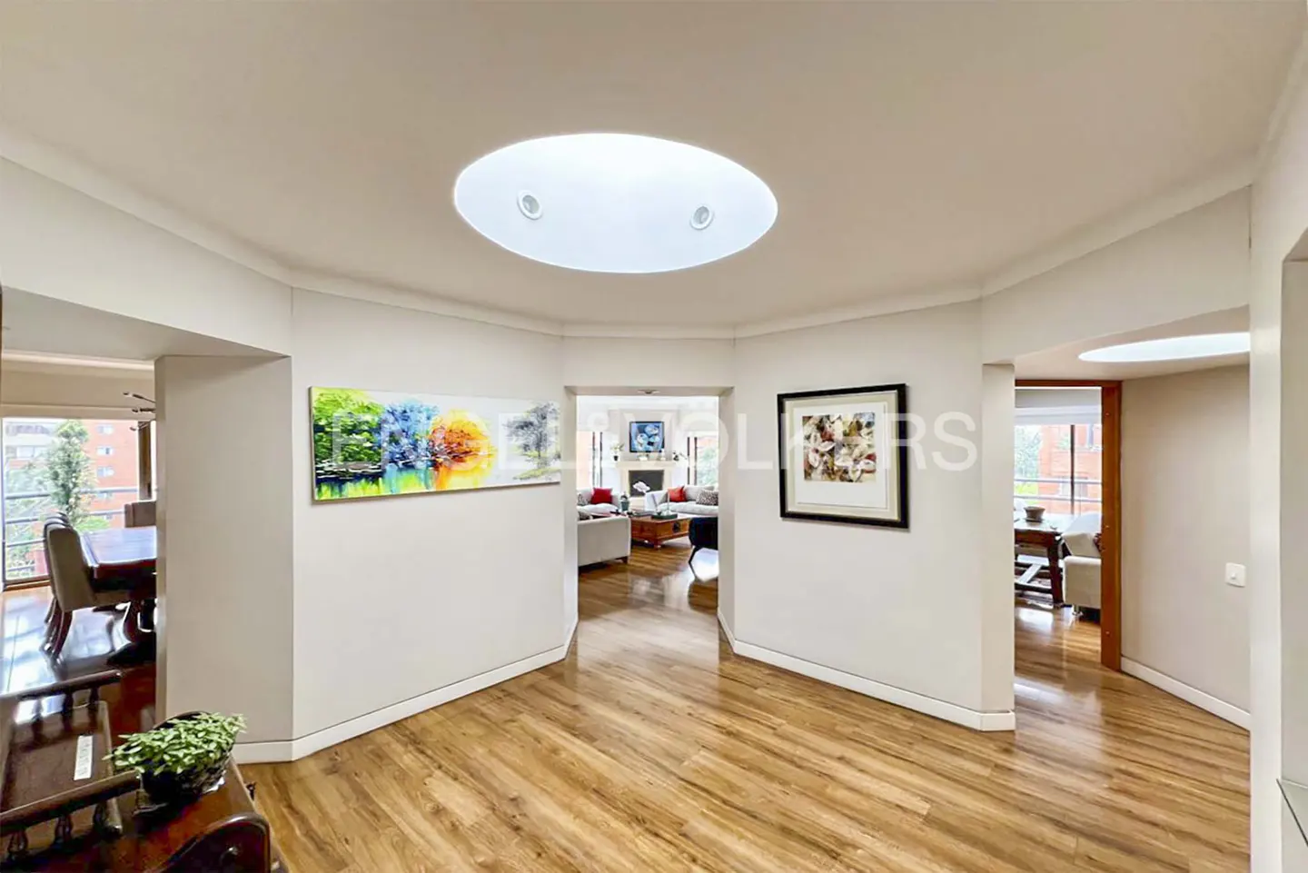 A bright foyer with wood floors, white walls, and a skylight. Artwork hangs on the walls. Open doorways lead to a dining room and living room.