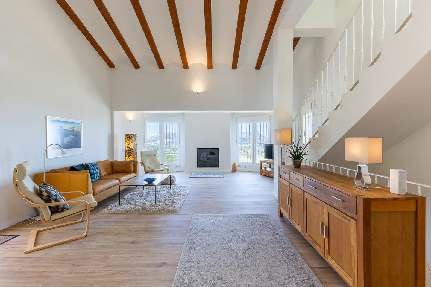Bright living room with white walls, wood floors, and exposed beams. A tan leather sofa and wooden cabinet furnish the space.