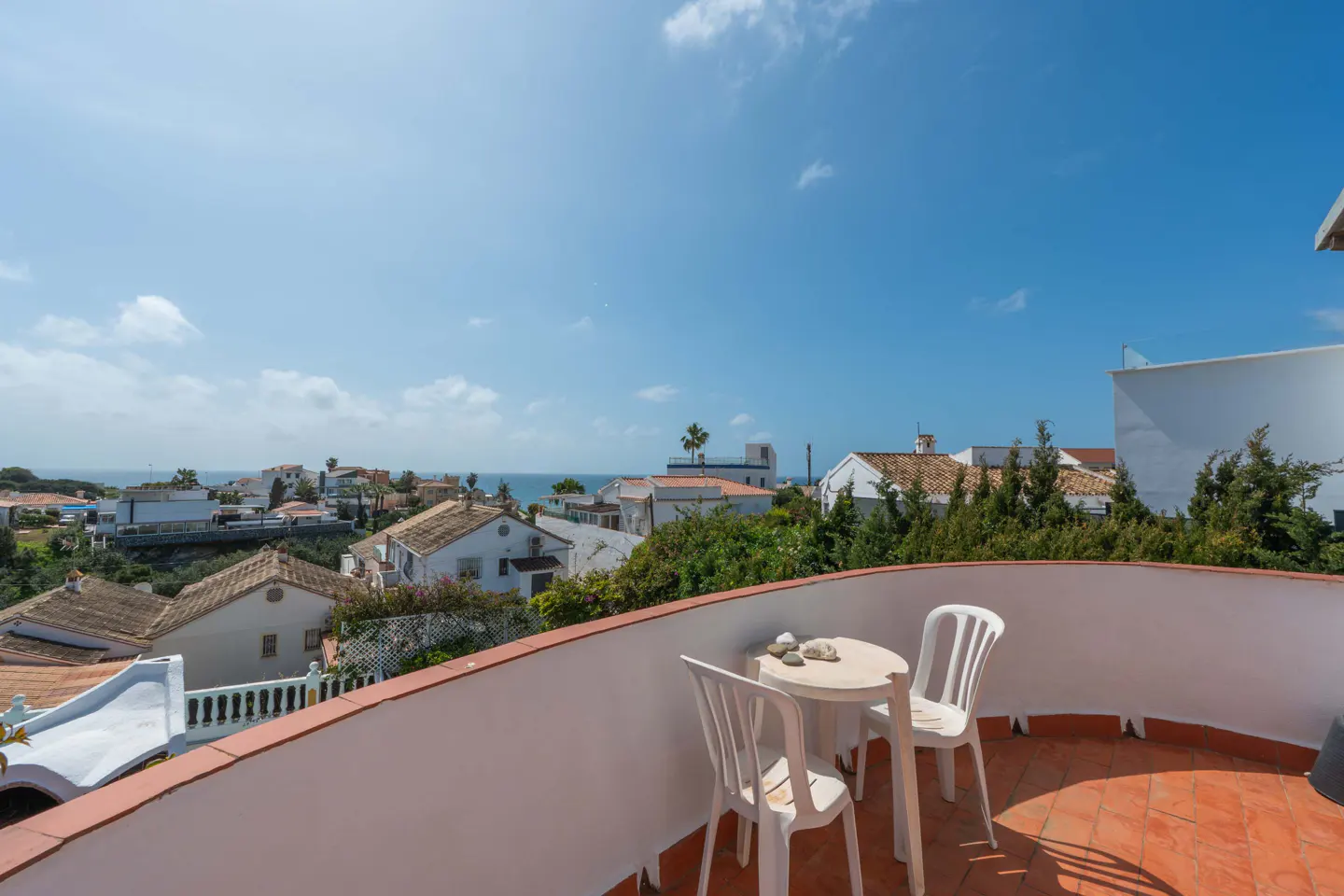 Balcony view with white table and chairs overlooking a coastal town and the ocean under a blue sky.