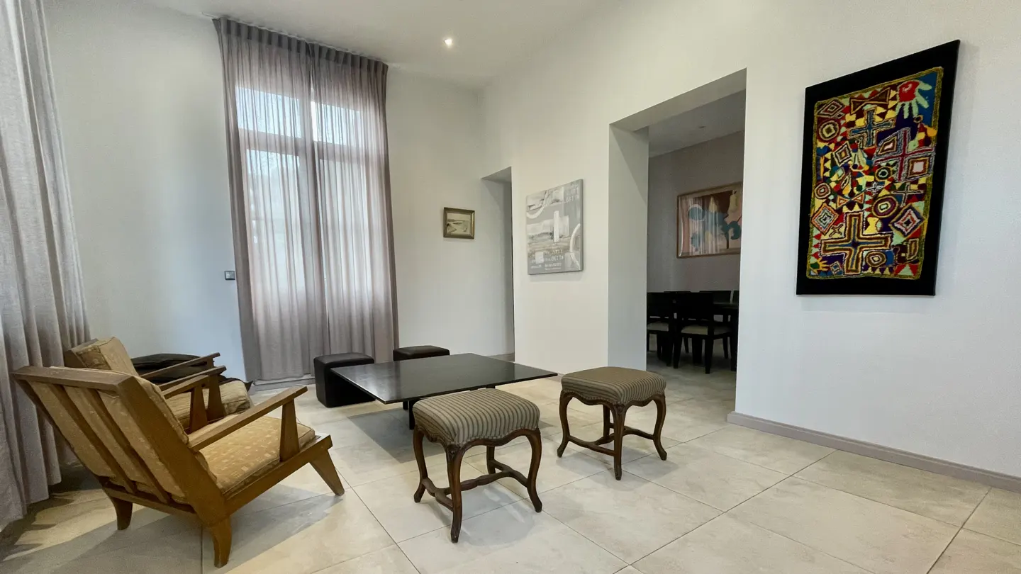 Living room with beige tile floor, white walls, wood chairs, black table, and colorful art.