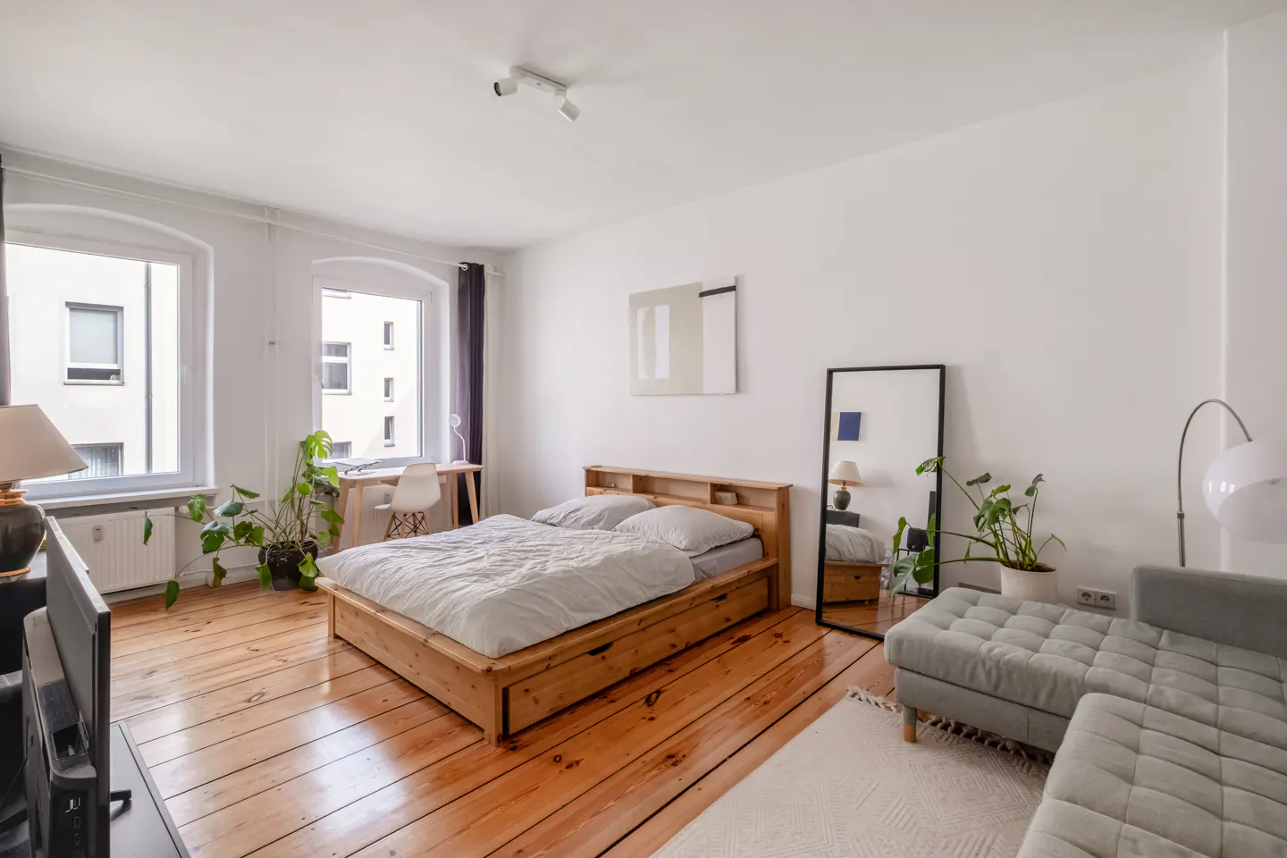 Bright bedroom with wood floors, a wooden bed with gray linens, a gray sofa, and large windows. A desk and plants add a touch of nature.