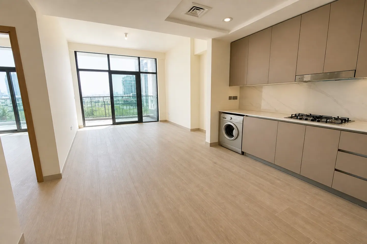 Bright, empty apartment with wood floors, beige cabinets, and a washing machine. A large window overlooks a green landscape.