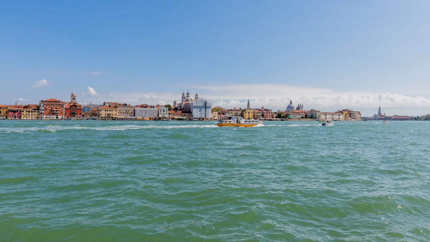 A yellow water taxi cruises across the turquoise Venetian lagoon with the historic city skyline in the background.