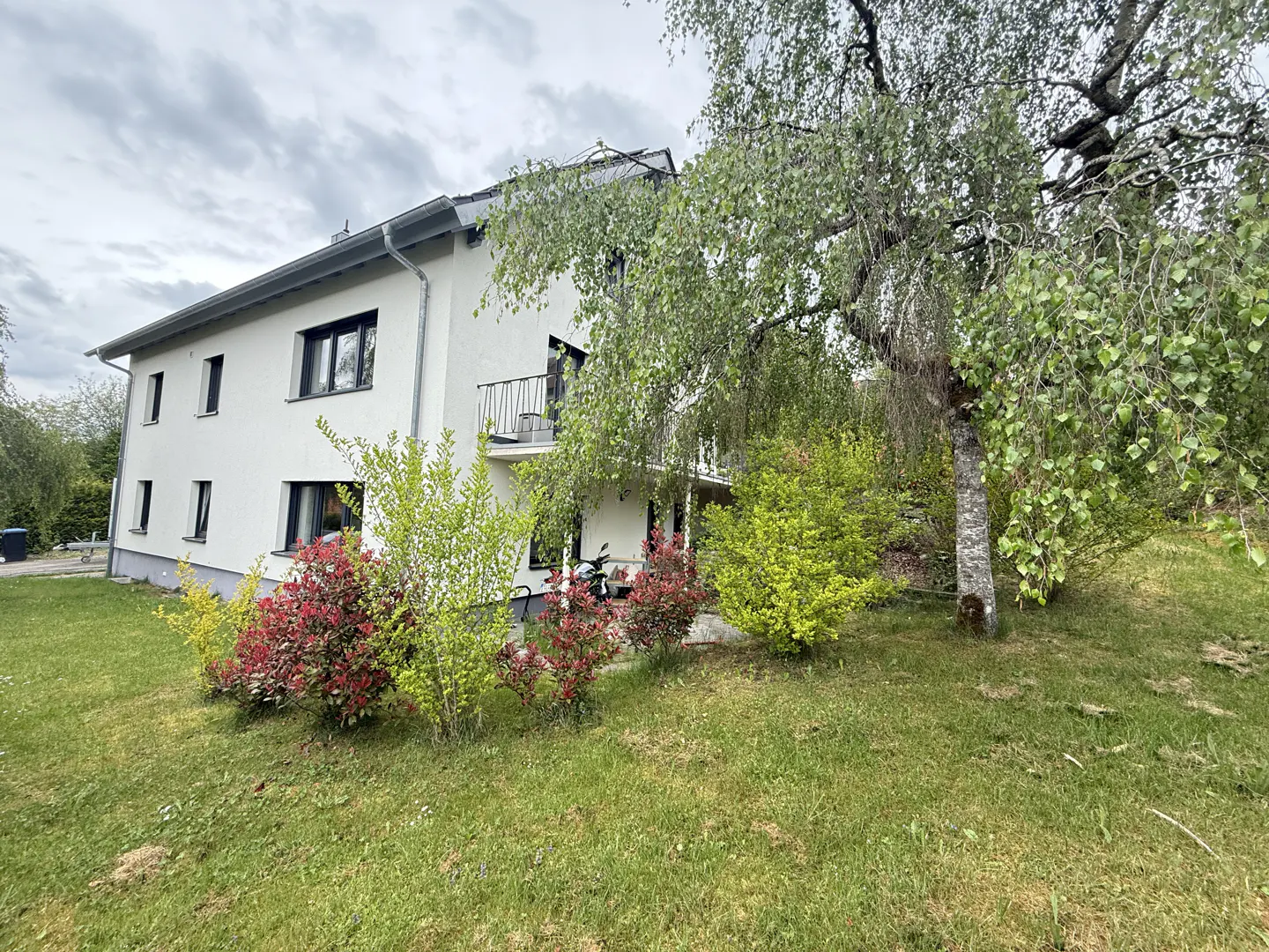 Two-story white house with a gray roof, black windows, and a small balcony, surrounded by green grass, bushes, and a large tree.