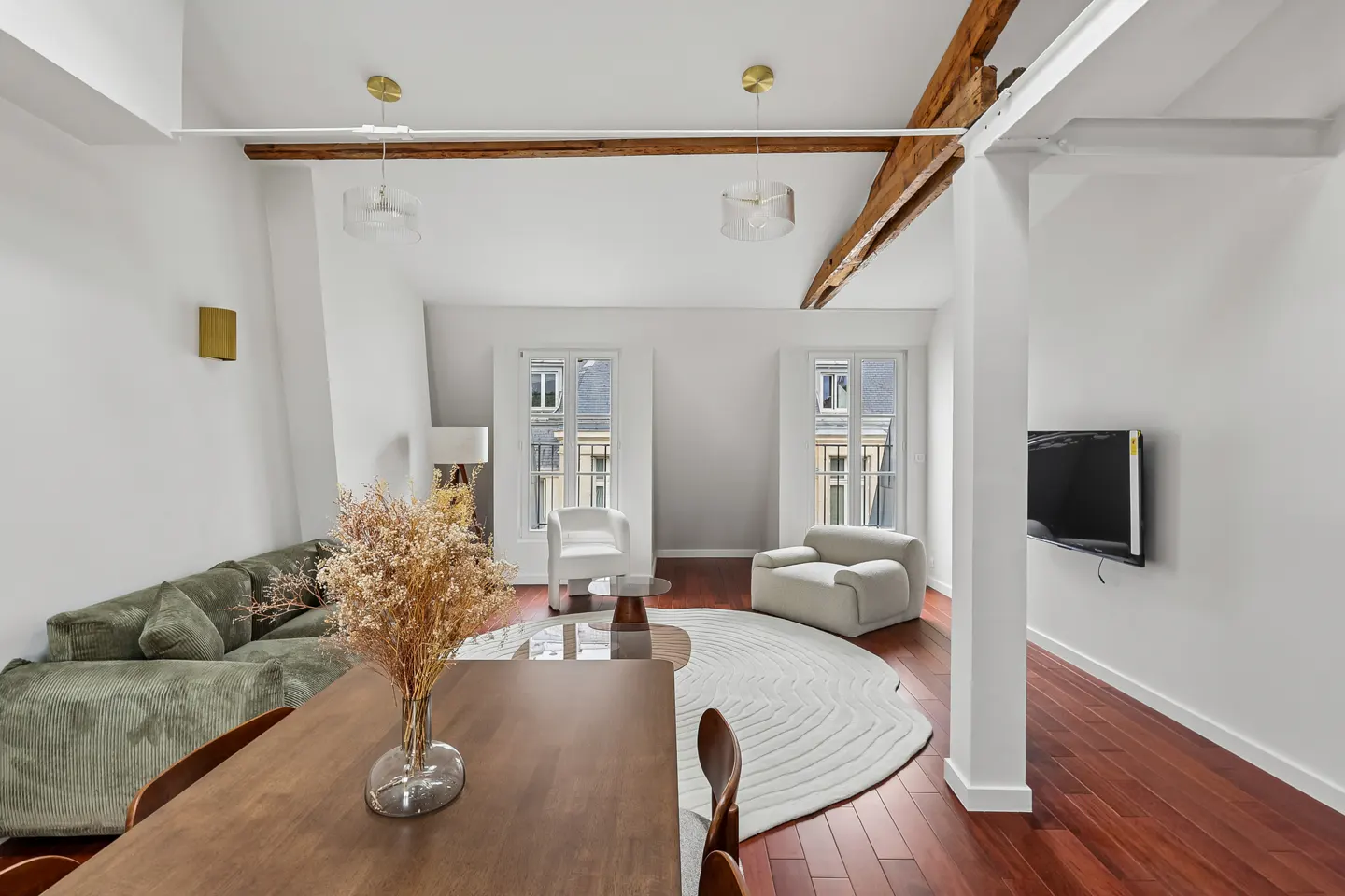 Modern living room with a green sofa, wooden dining table, white armchairs, round rug, and exposed wooden ceiling beams.