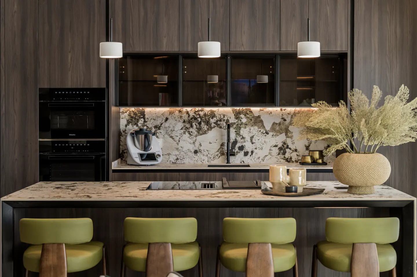 A modern kitchen with dark wood cabinets, marble countertops, and four green bar stools. Three white pendant lights hang above the island.
