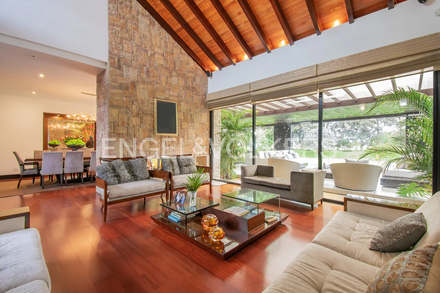 A spacious living room with a stone wall, wood ceiling, and large windows overlooking a garden. Sofas and a glass coffee table furnish the room.